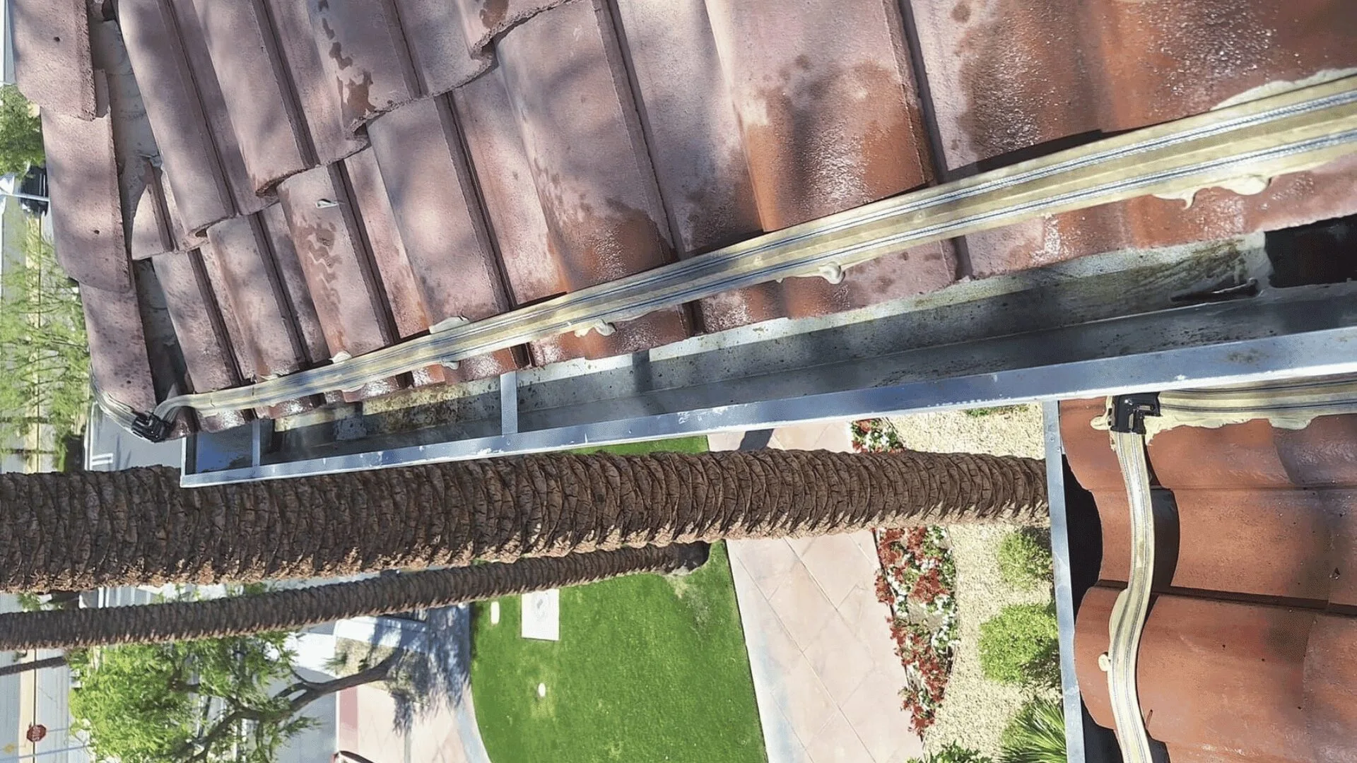 Overhead view of a red clay-tiled roof edge with a metal gutter and sealing compound along the eaves.