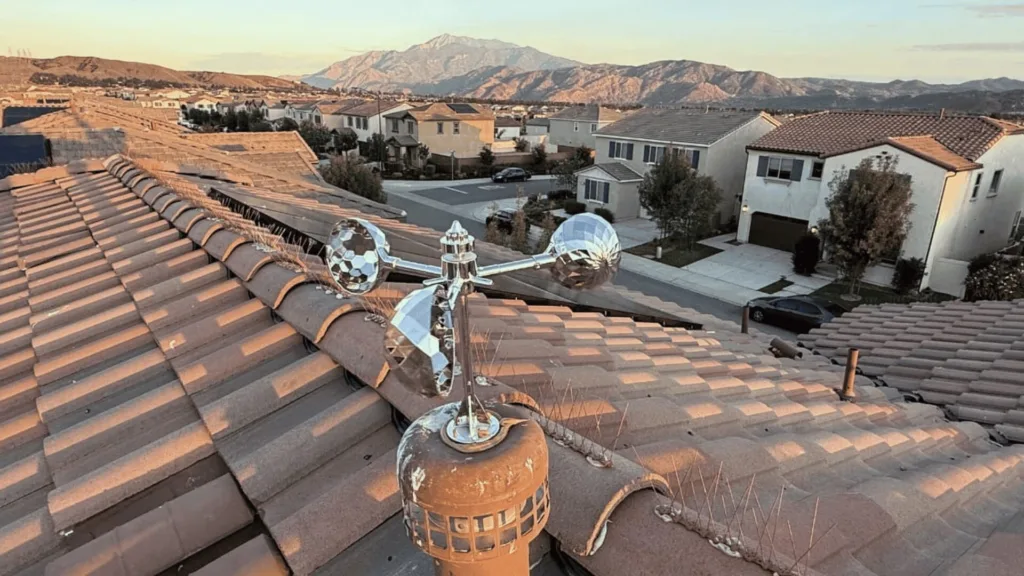 Shiny chrome weather vane with three mirrored globes mounted on a tile roof overlooking a suburban street and distant mountains at sunset.