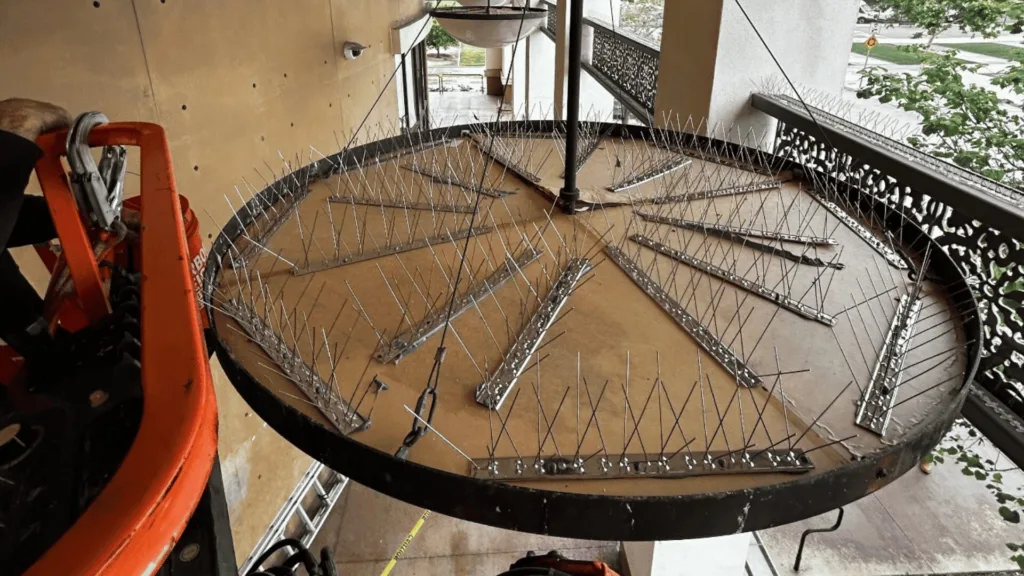 Circular metal platform covered in evenly spaced vertical spikes arranged in spokes around a central pole, on an outdoor balcony with construction equipment nearby.