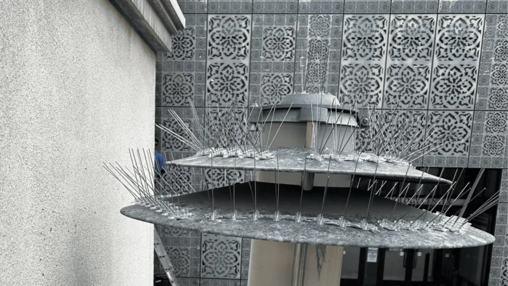 Circular metal platform with numerous long spikes around the rim, outdoors against a patterned wall with decorative panels.