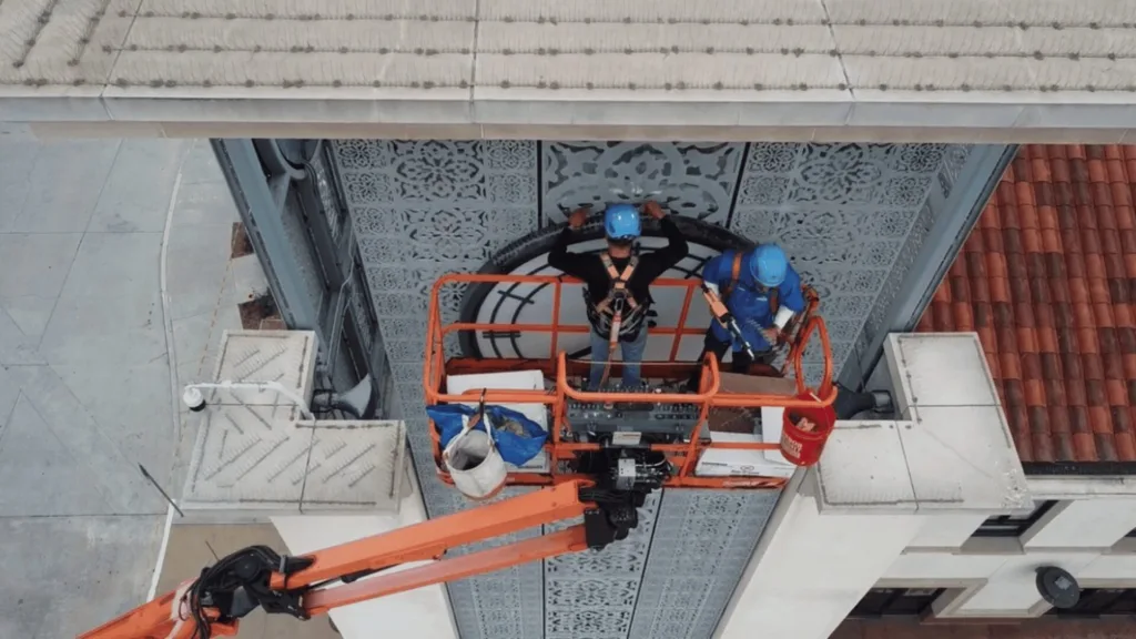 Two workers in hard hats on an orange suspended lift repairing decorative metal facade on a tall building
