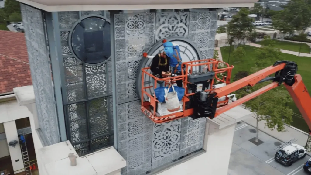 Two workers in blue uniforms on an orange aerial lift performing exterior maintenance on a decorative metal facade with circular and floral patterns.