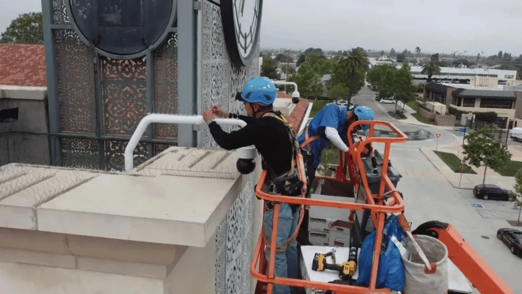 Workers in blue helmets on a lift installing or inspecting a railing on a building rooftop edge.