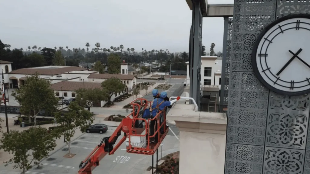 Two workers in blue uniforms on an elevated platform inspecting a decorative clock tower beside a city street.