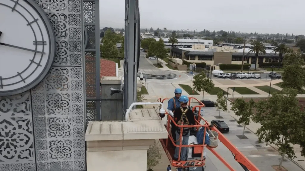 Two workers wearing blue helmets on a red elevated lift beside a clock-tower with decorative metal paneling; urban street below in the distance.
