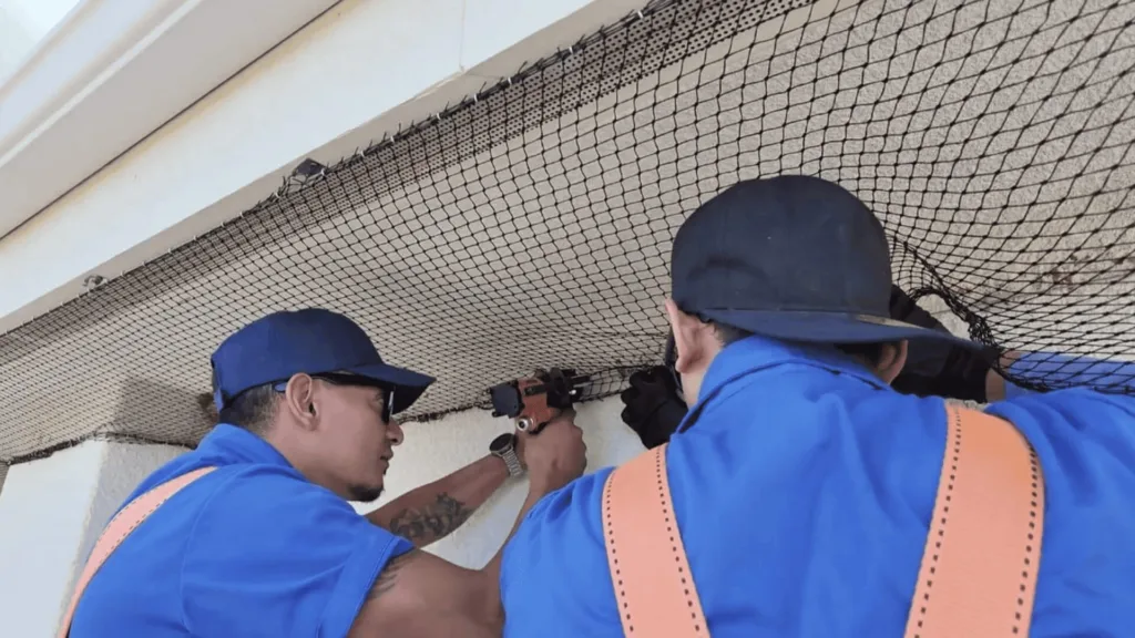 Workers in blue uniforms installing a black safety net along a ceiling, using tools at a work site.