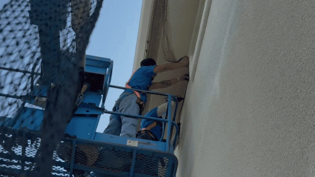Worker in a blue shirt wearing a safety harness on a raised blue lift, reaching into a window opening of a beige building exterior.