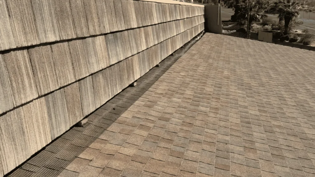 Rooftop scene showing textured shingle siding along the left and a sloped shingled roof extending to the right, with trees and a road in the distance.