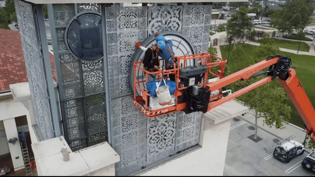 Two workers on an orange aerial lift cleaning/repairing a decorative metal facade on a tall building.