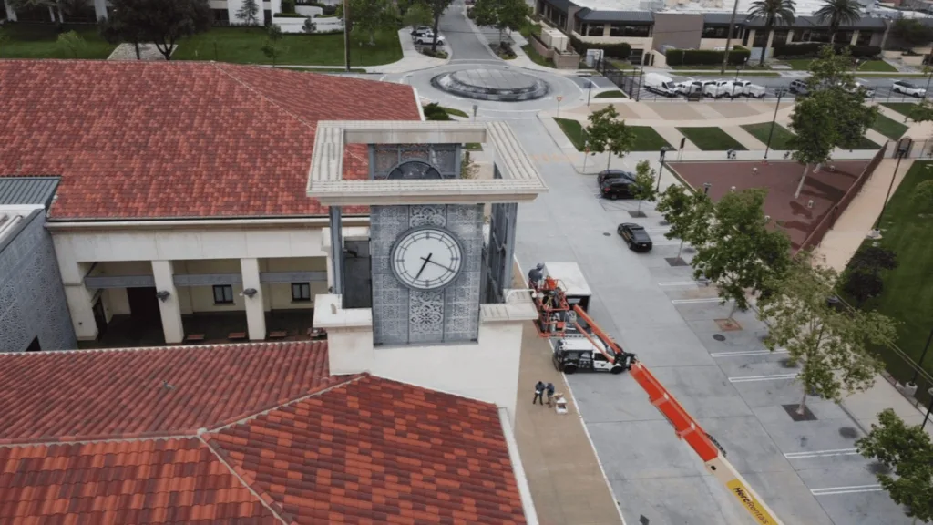 Aerial view of a clock tower on a campus building with a red-tiled roof; a construction lift is attached to the tower and workers are nearby.