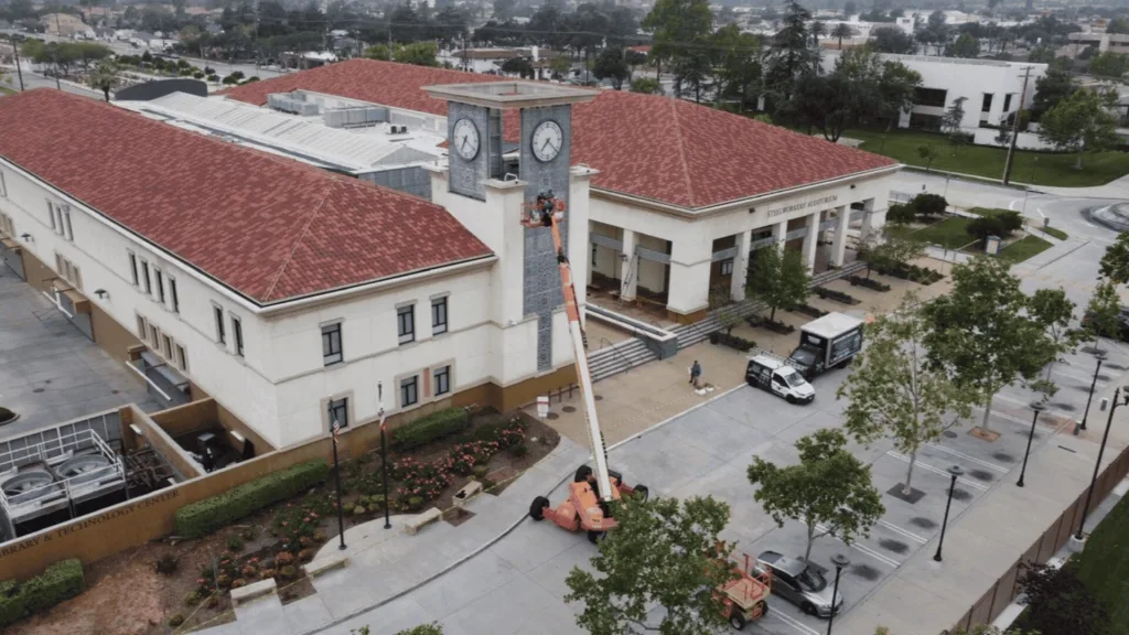 Aerial view of a campus building with red-tiled roof and a clock tower; a cherry picker lifts a worker to the clock faces on the tower.