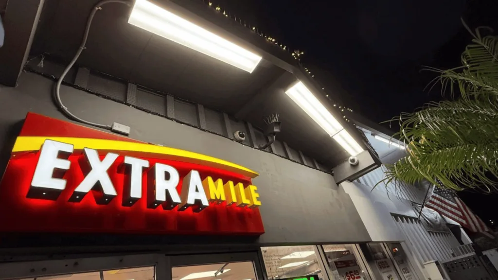 Nighttime exterior of a building with a bold red and yellow sign reading EXTRAMITE above a windowed entrance, string lights along the roofline, and palm fronds to the right side.