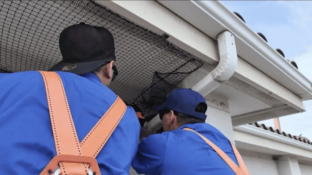 Two workers in blue shirts and orange safety harnesses install a mesh gutter guard on a house exterior under a blue sky.