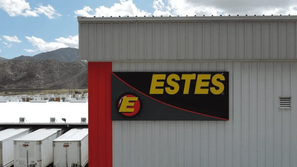 Grey corrugated warehouse with a large Estes sign (yellow letters) and a red vertical accent; white trailers lined up in front against a mountainous backdrop.