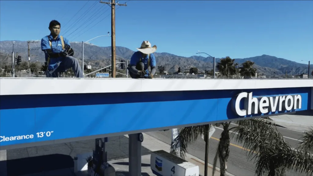 Two workers in blue uniforms stand on a Chevron billboard structure against a clear blue sky and distant mountains.