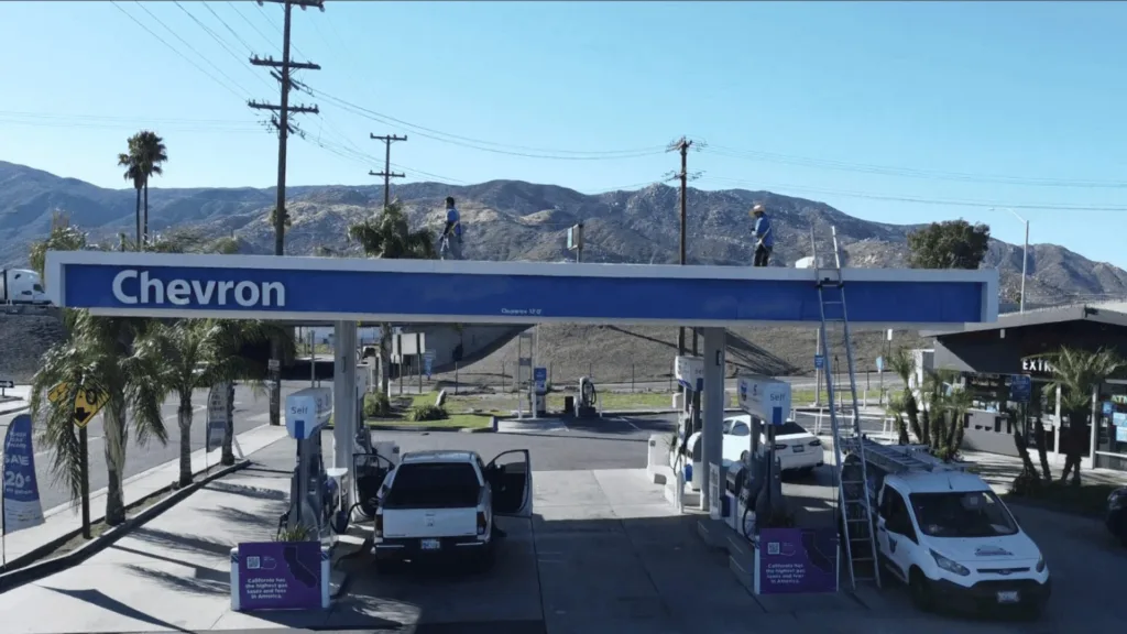 Chevron gas station with blue canopy and several white vans refueling side by side, hills in the background