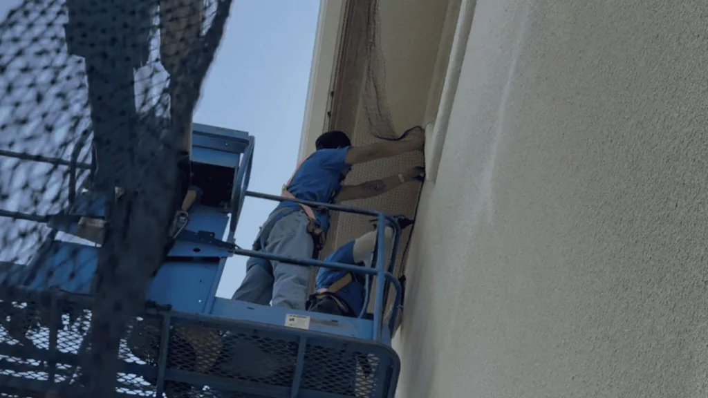 Two workers in blue shirts on a blue hydraulic lift against a building wall, one reaching into a window or opening guarded by netting nearby.