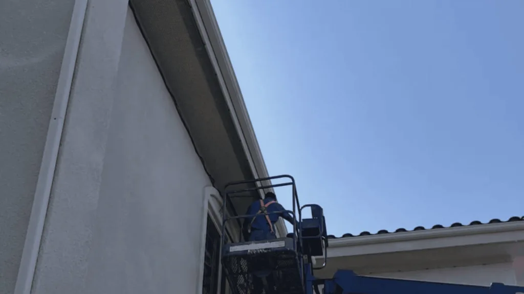 Maintenance worker in a safety harness on an elevated platform next to a curved roof of a building against a clear blue sky.