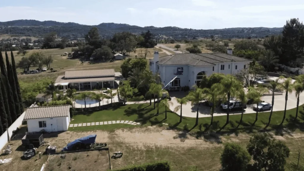 Aerial view of a large white villa with tiled roof, palm trees, and a circular driveway, set among green lawns and hills in the distance.