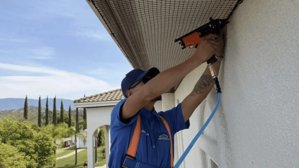 Worker in blue uniform using a pneumatic nail gun to fasten material to an exterior wall under the eave, with a blue air hose attached.