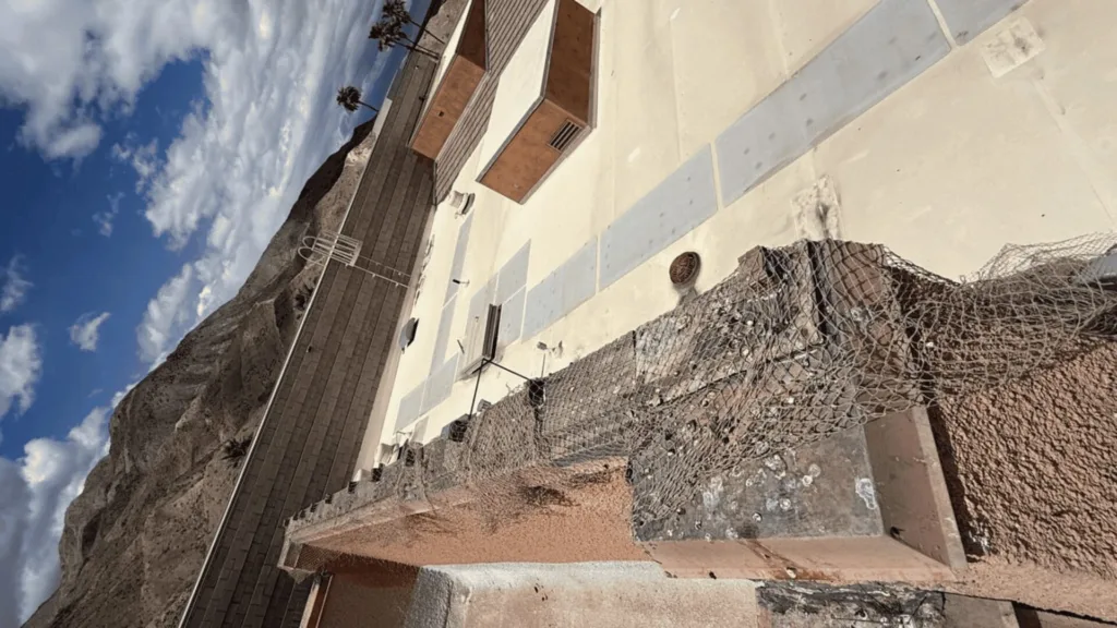 Concrete terrace along a rocky cliff with a metal railing and weathered chain-link barrier under a blue sky with clouds.