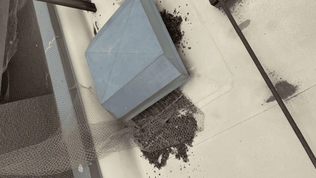 Maintenance rooftop scene with a gray metal exhaust vent amid debris and protective netting on a concrete surface