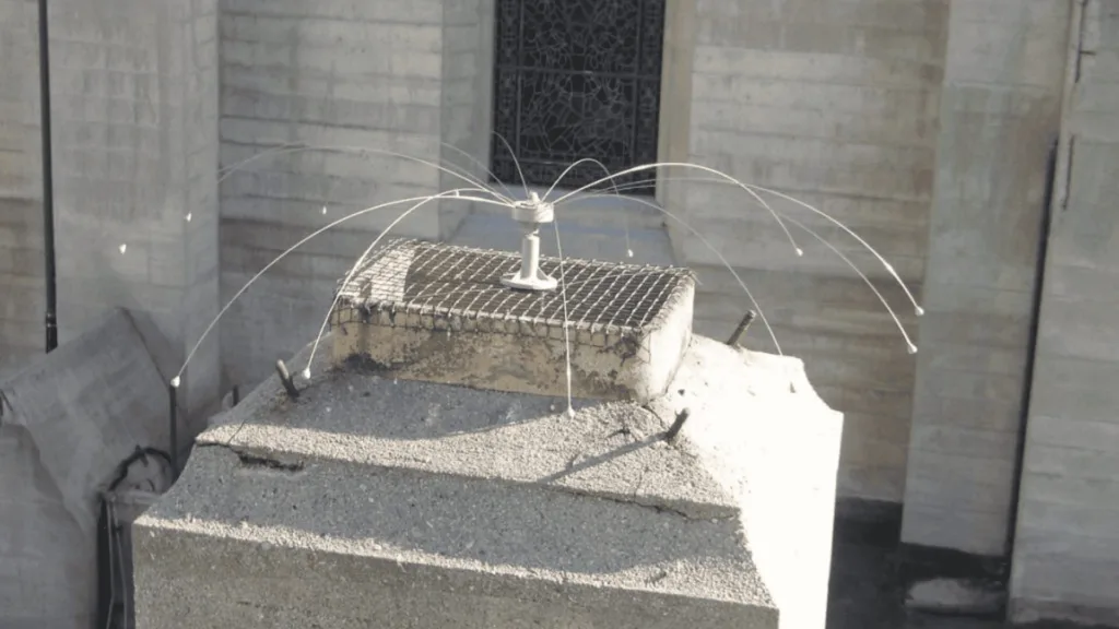 Industrial rooftop sprinkler with multiple jets arching outward from a central hub on a concrete block, spraying water in arcs.