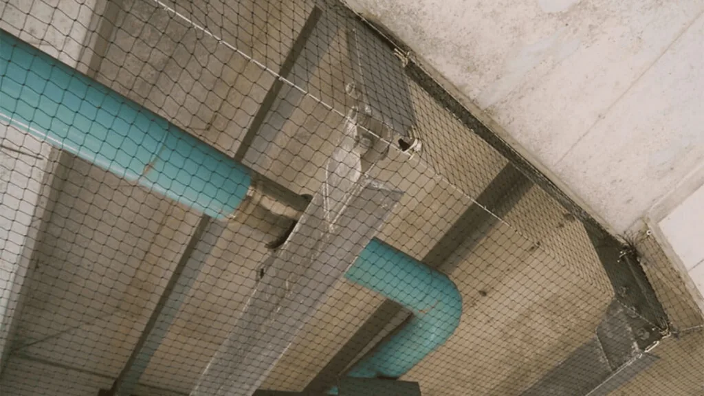 Interior industrial space with turquoise ductwork running along a concrete ceiling behind a chain-link fence in a dimly lit area, viewed from below.
