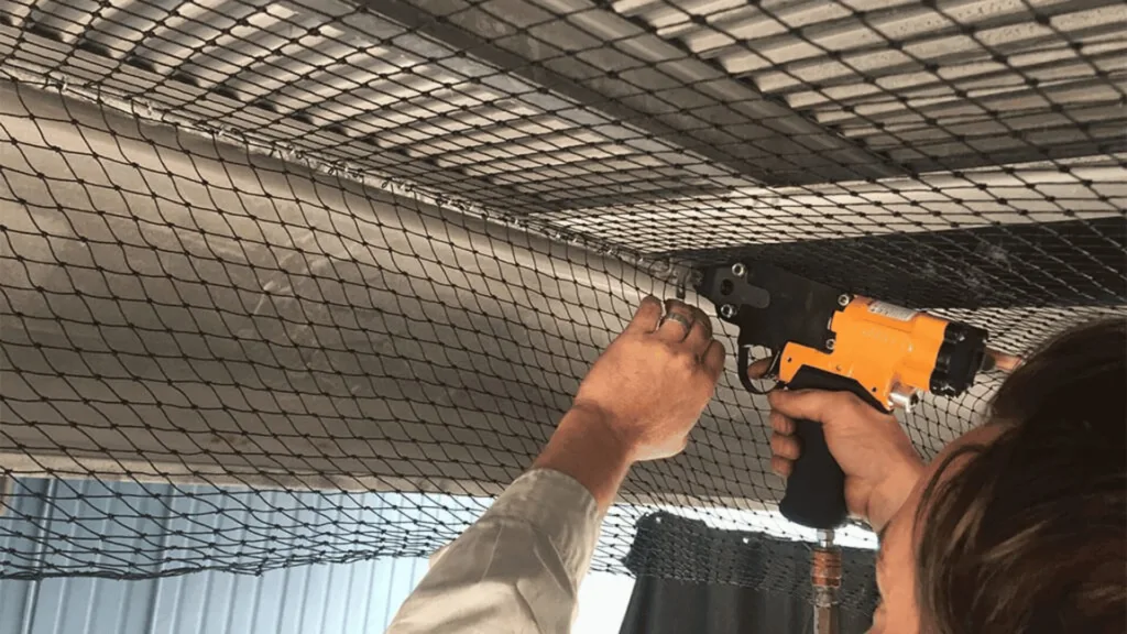 Worker uses an orange pneumatic nail gun to attach metal mesh to a ceiling grid.