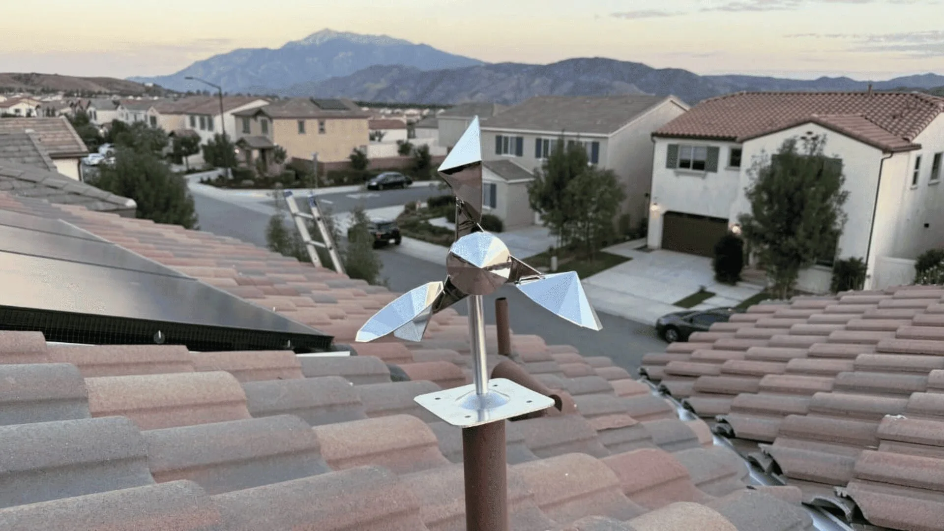 Shiny geometric wind vane mounted on a brown pole atop a tiled roof, with a suburban street and mountains in the distance.