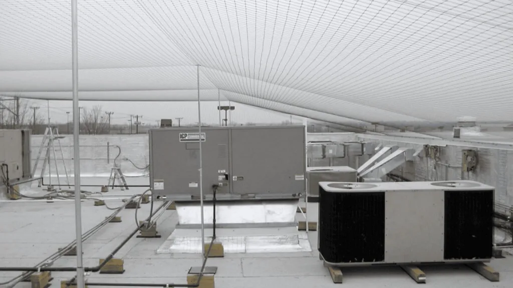 Rooftop HVAC equipment on a flat roof with large air handlers and wiring under a mesh shade structure in a cloudy setting.
