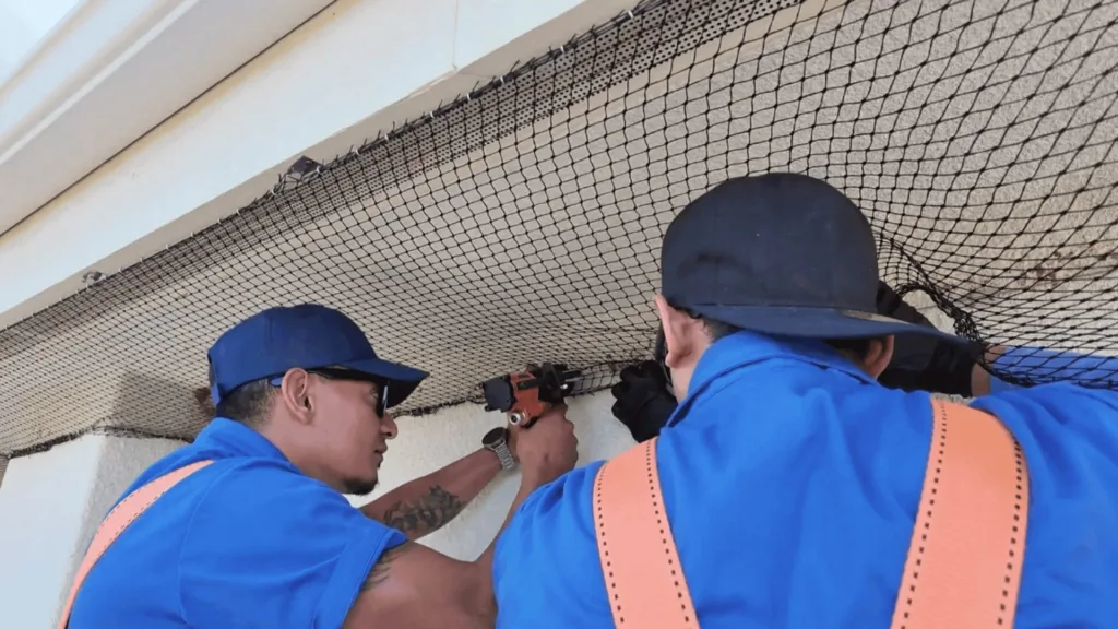 Three construction workers in blue uniforms install a black safety net along a curved ceiling edge.