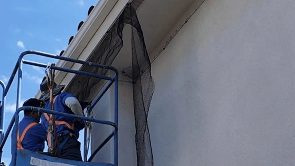Two construction workers in safety harnesses on a blue elevated platform repairing a building facade with protective netting nearby.