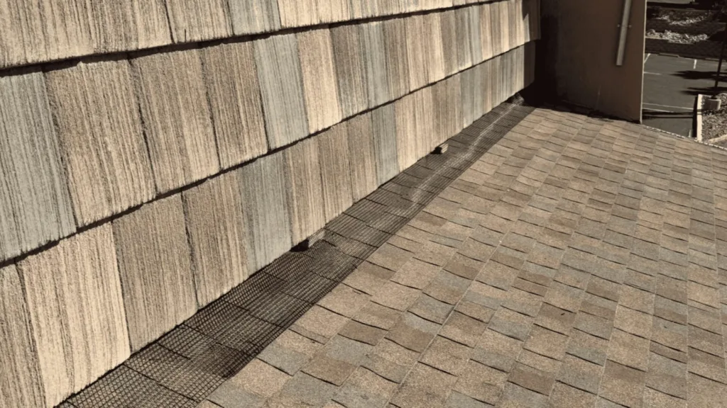 Outdoor walkway beside a textured, stacked-stone wall with a metal drainage grate along the base edge, and a small dark object on the ground nearby.