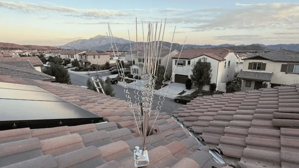 Rooftop antenna with multiple metal rods rising above terracotta tiles in a suburban neighborhood with mountains in the distance