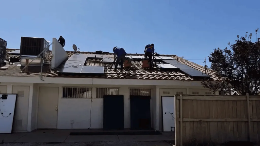 Workers on a tiled roof installing solar panels under a clear blue sky.