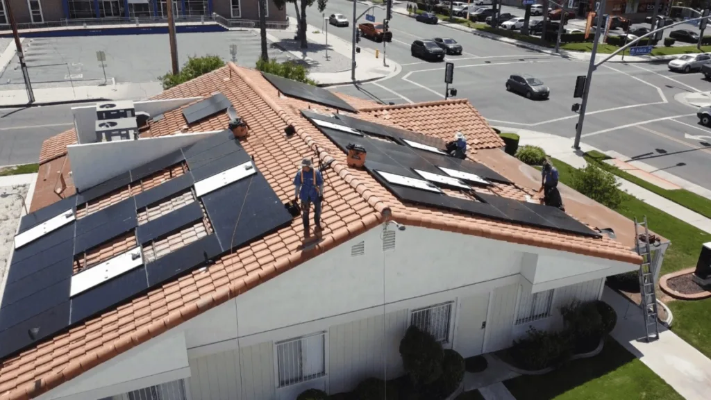 Workers install solar panels on a terracotta-tiled roof above a white building near a busy intersection.