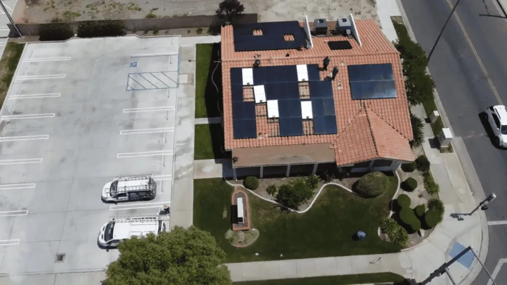Aerial view of a red-tiled roof building covered with dark solar panels, with a large parking lot to the left and a street to the right.
