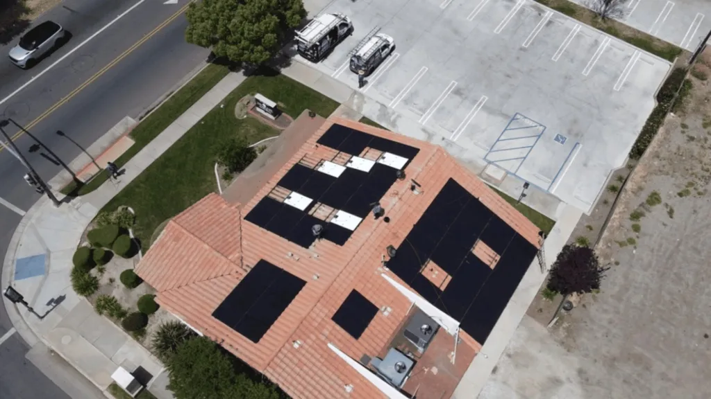 Aerial view of a building with solar panels on a red-tiled roof beside a parking lot and street, showing cars and trees nearby.