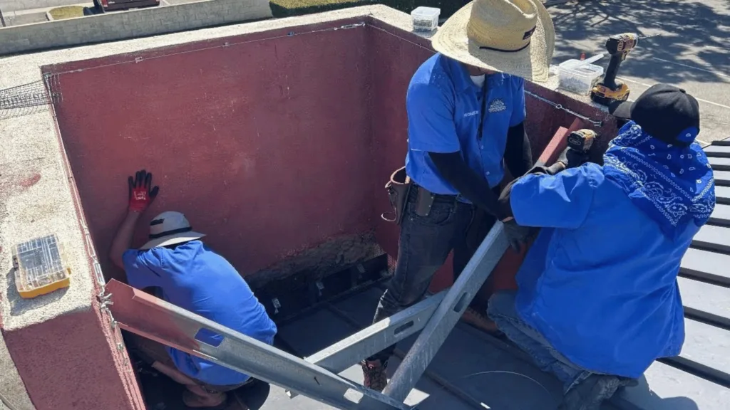 Three construction workers in blue shirts on a rooftop install a metal ladder into a red open hatch, with tools nearby.
