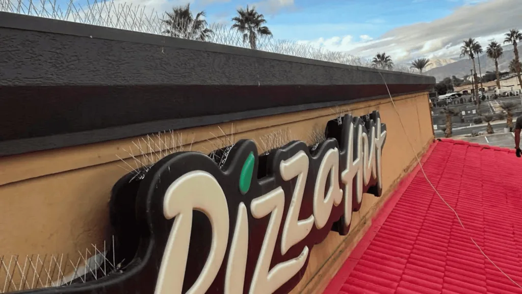 Part of a large 'Pizza' sign on a beige wall above a red roof, with barbed wire along the top and palm trees in the background.