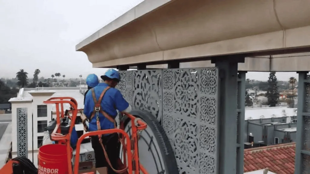 Worker in blue safety gear on a lift, handling equipment on a rooftop next to decorative metal screens and a concrete overhang