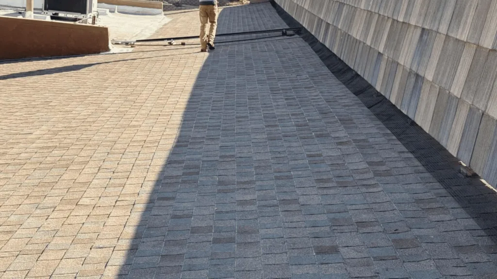 A person walking away on a rooftop with tan-paved tiles, split into light and dark sections and a tall sloped wall on the right.