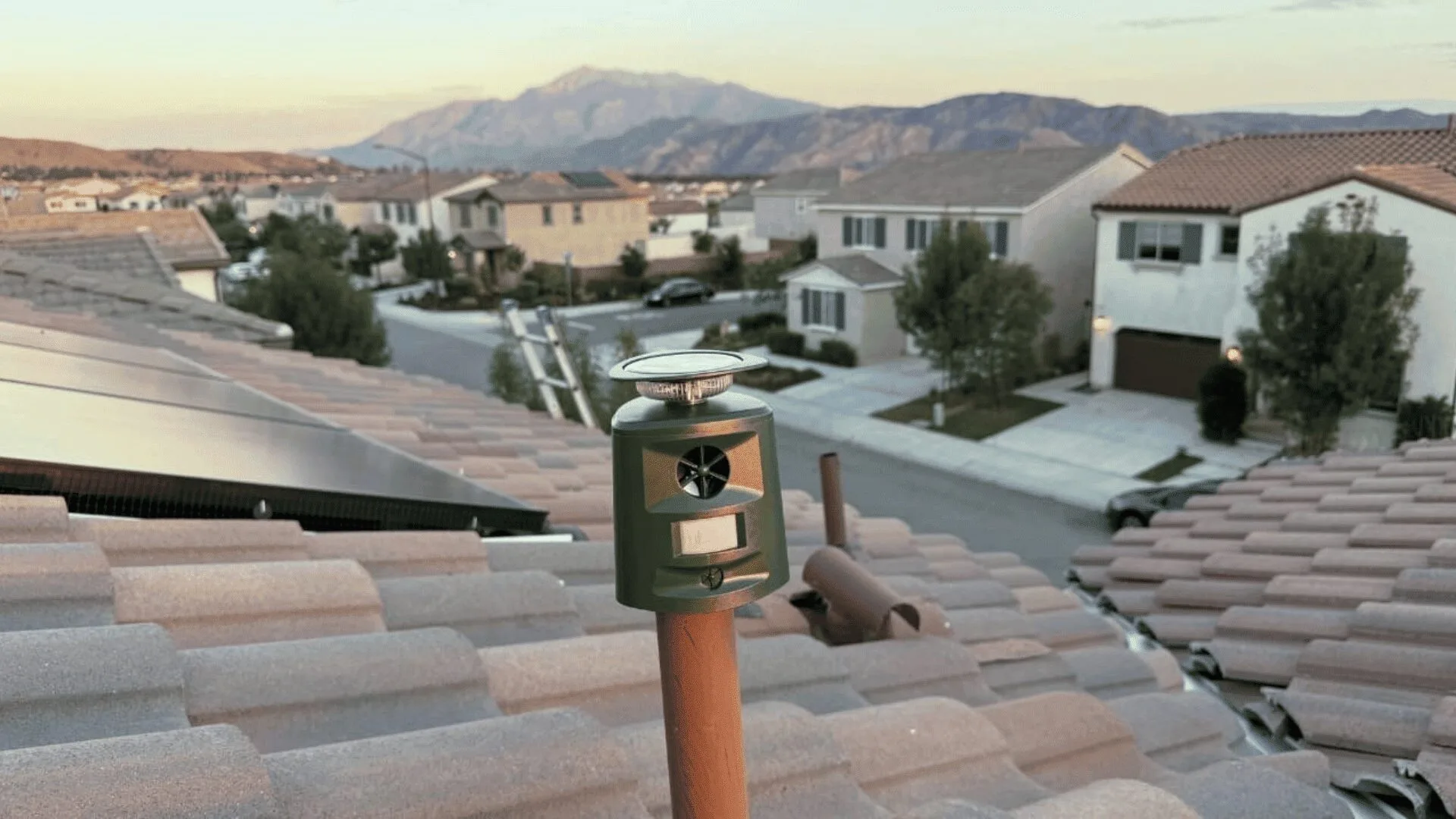 Green roof-mounted turbine vent on a brown vent pipe, overlooking a suburban street with tile roofs and distant mountains (attic ventilation device).