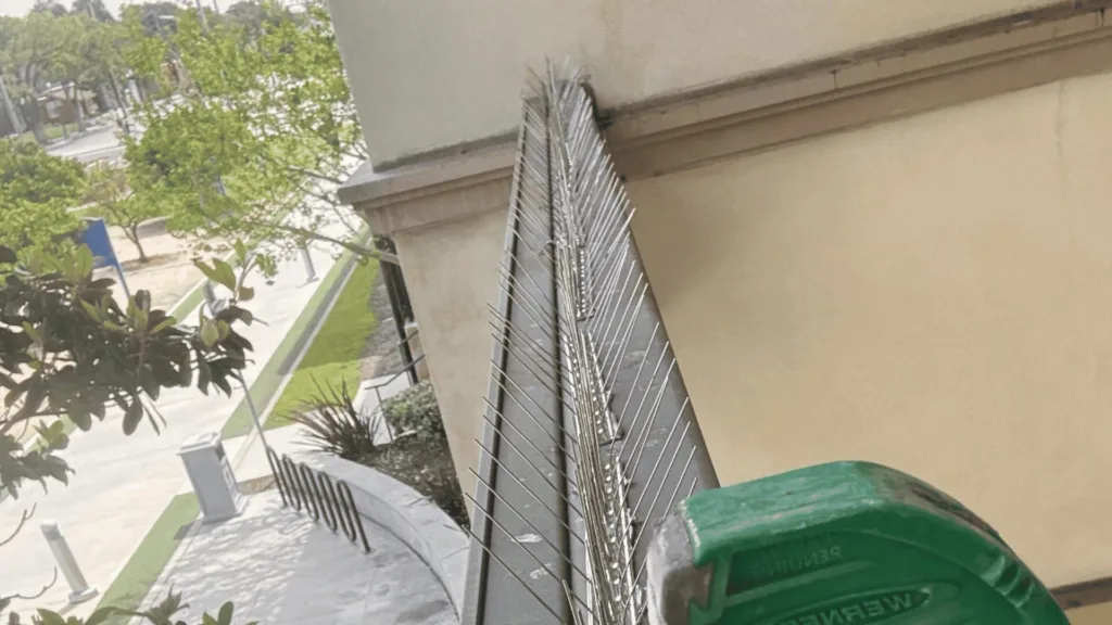 Tall, spiked metal security fence mounted along a beige building, with a green trash bin in the foreground and trees in the background.
