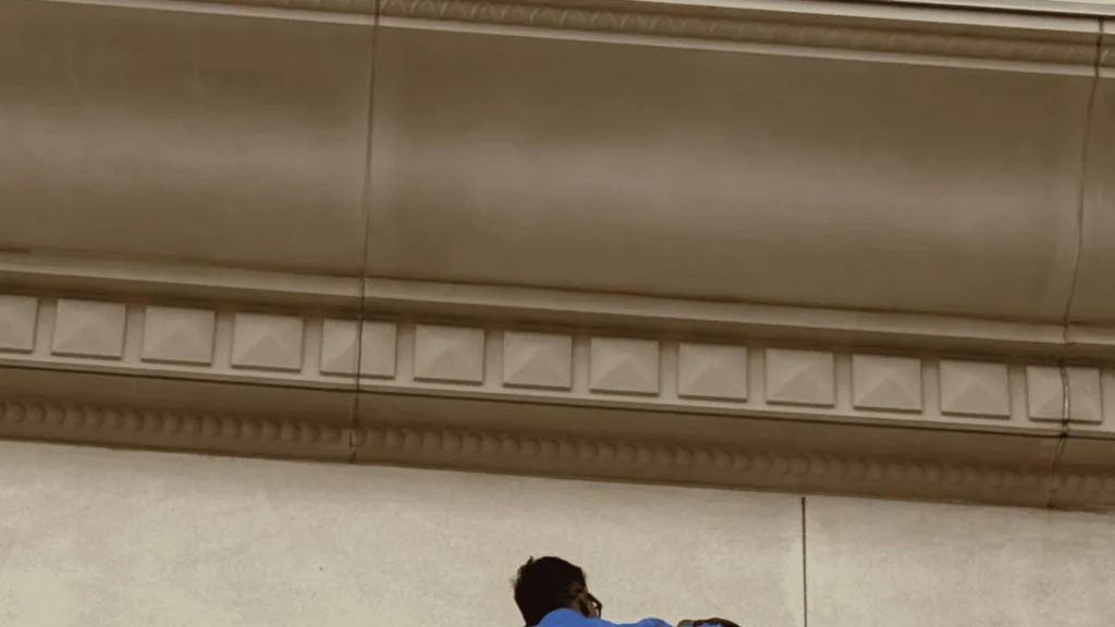 Close-up view of an ornate ceiling with square decorative panels and molding; a person in blue appears at the bottom edge.