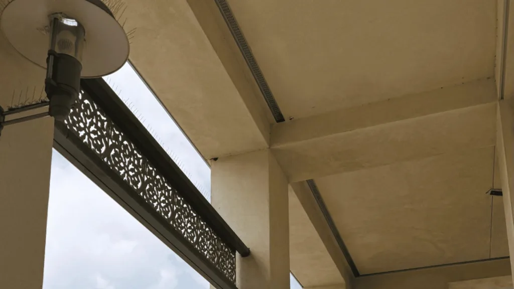 Beige concrete balcony area with a round wall light and decorative lattice railing by a large window.