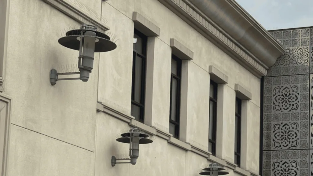 Exterior view of a beige building wall with wall-mounted cylindrical lamps and rectangular windows, plus a decorative patterned panel on the right.