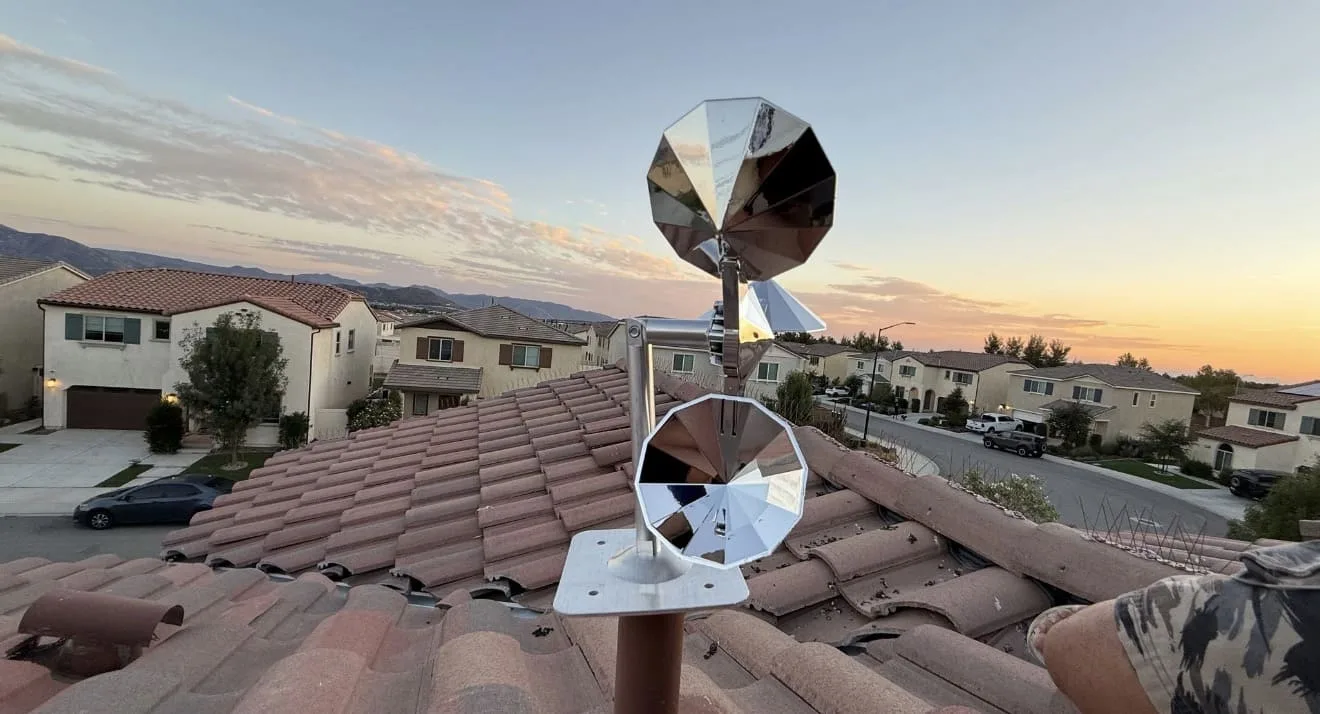Shiny metallic wind sculptures on a tiled rooftop overlooking a suburban neighborhood at sunset.