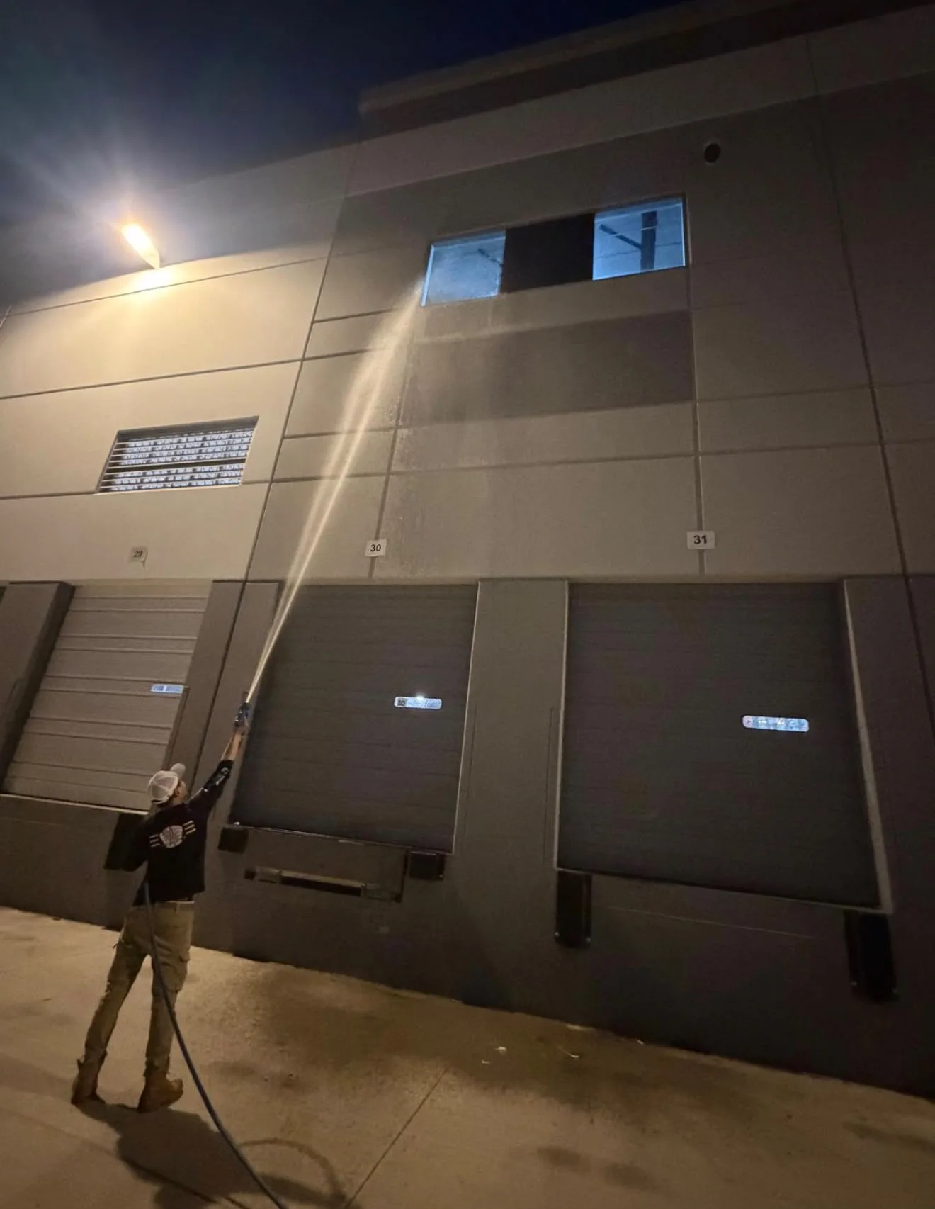 Person operating a high-pressure hose washing the exterior wall of a gray industrial building at night.